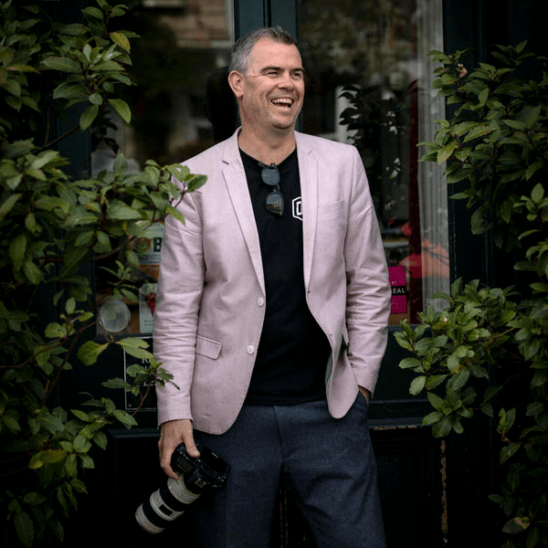 A portrait of professional wedding photographer John Sanders holding a camera stood outside a bar in London.